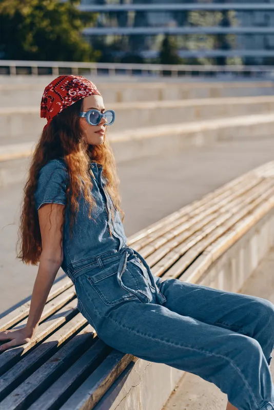 young woman wearing denim outfit and red bandana sitting on wooden bench outdoors in sunny weather with sunglasses and long curly hair