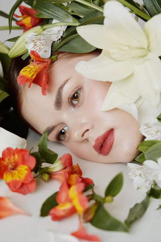 Young woman with natural makeup surrounded by fresh flowers, expressing tranquility and beauty in a serene composition featuring vibrant colors