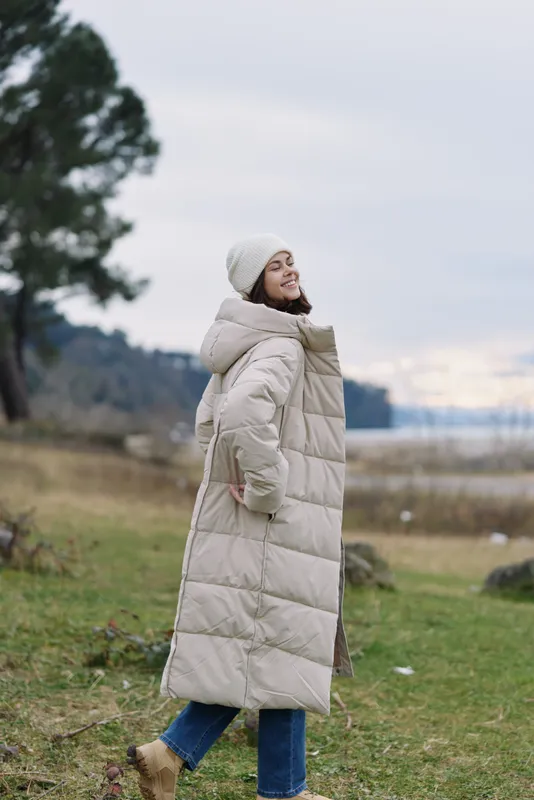 Woman wearing a long beige puffer coat in an open outdoor landscape, smiling with hands in pockets, casual winter fashion amid tranquil countryside and lake.