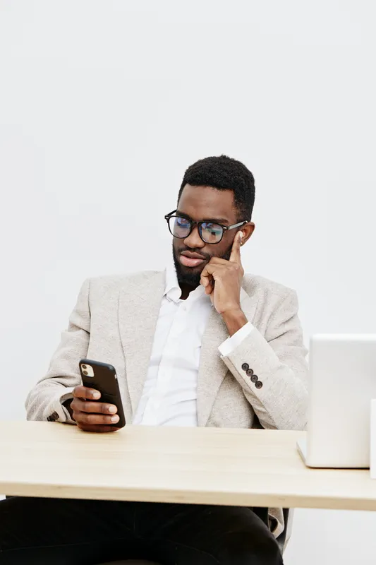 professional man using smartphone wearing glasses and formal jacket sitting at desk in office with laptop isolated on plain white background studio shot. Business workplace concept
