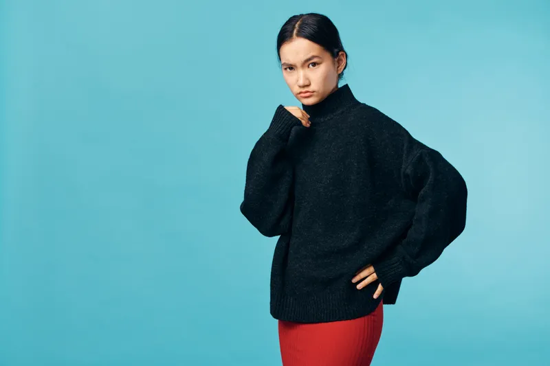Young woman with dark hair wearing black sweater and red skirt standing against blue background looking serious with thoughtful expression and hand near chin.