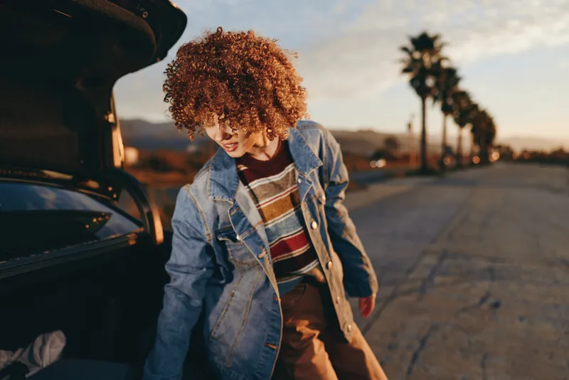 Woman lifestyle, curly hair woman in denim jacket leans near open car on quiet street at sunset, warm golden light, relaxed mood, urban palms and road perspective, evening trip alone