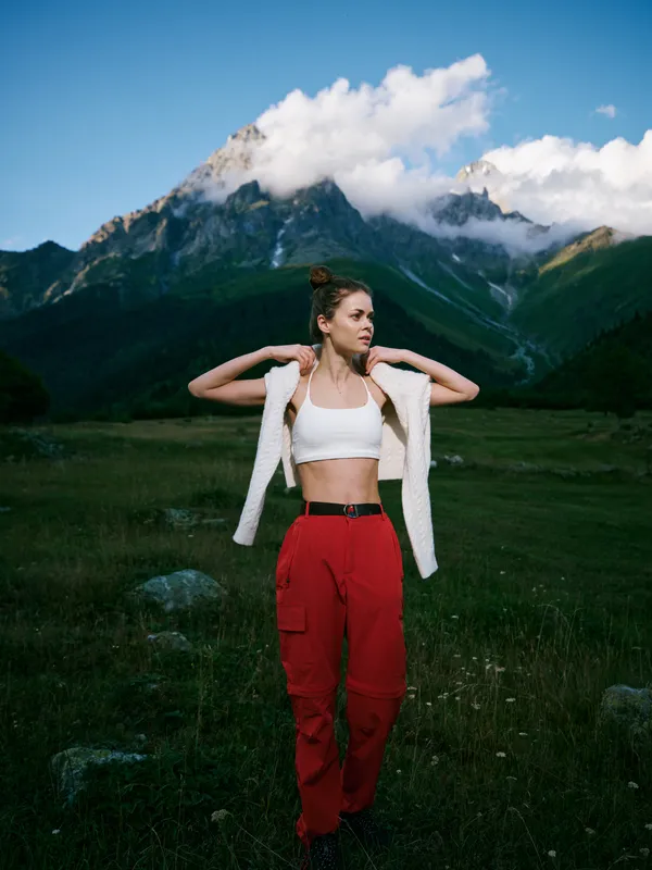 Outdoor landscape with a woman wearing a white crop top and red cargo pants, standing in a grassy valley as rugged mountains rise under a blue, cloudy sky