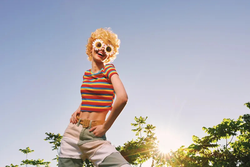 Cheerful woman wearing a striped crop top and shorts, playful mask on the face, posing outdoors with bright sunlight, casual lifestyle scene, vibrant retro colors and carefree mood.