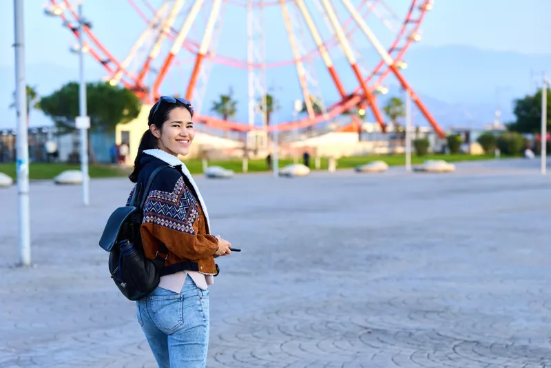 Young woman with a backpack at an amusement park, smiling beside a towering ferris wheel, casual jacket and jeans, outdoor travel mood, leisure photography capturing playful exploration and curiosity