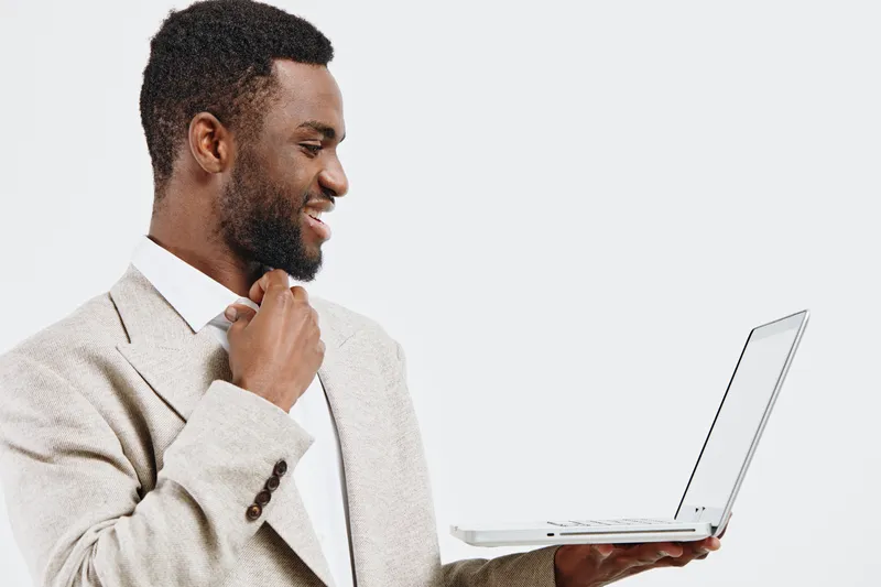 Professional young African American man wearing beige blazer and white shirt, holding a laptop in hand, smiling and looking at the screen, isolated on plain white background, studio portrait, business