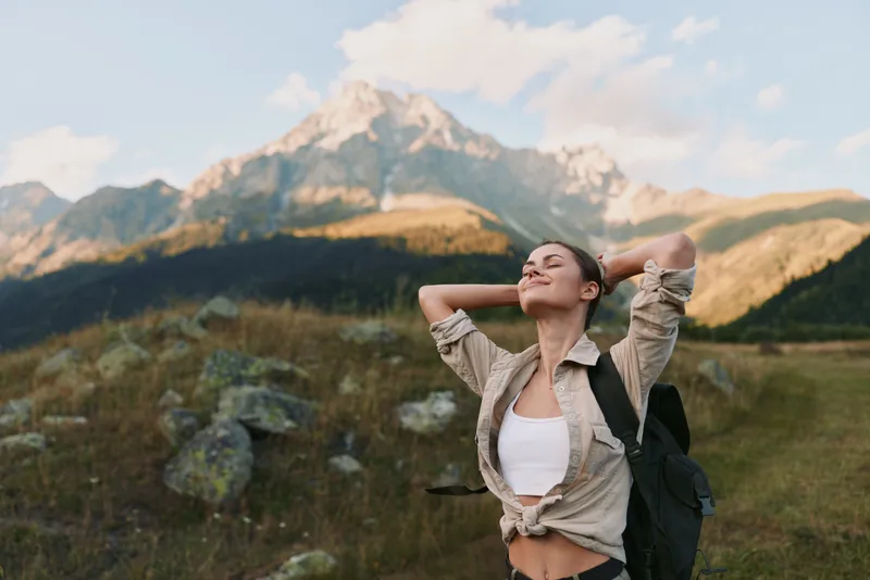 Woman outdoors, smiling with eyes closed, sunlit mountain landscape, relaxed posture, casual hiking attire, breeze in hair, nature colors, serene moment