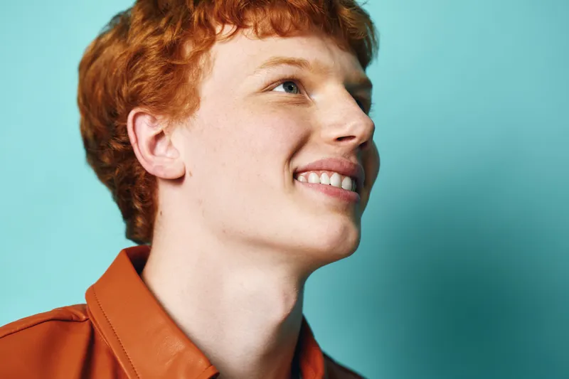 Young man with red hair smiling and looking up with teeth, wearing a brown shirt against blue background. Portrait, happiness, and positive emotion captured in studio.