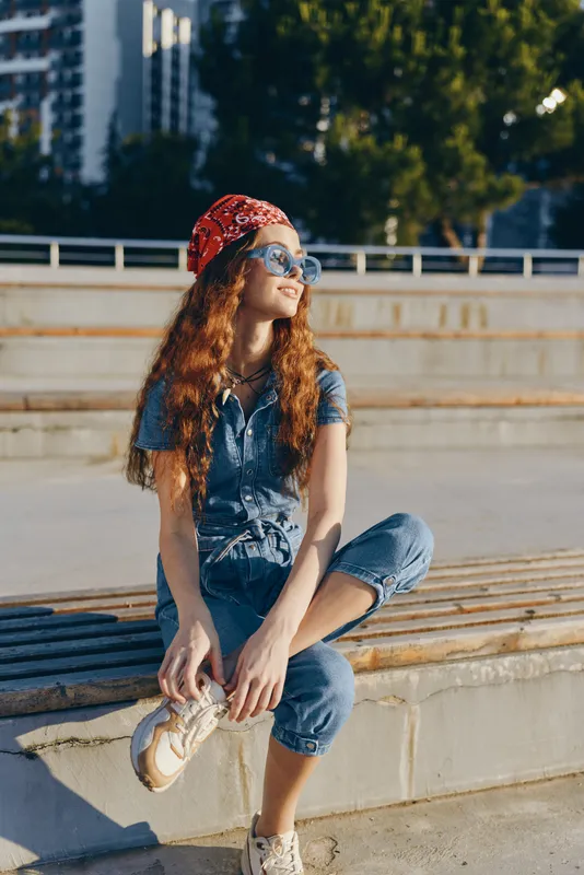 Lifestyle woman in modern boho-western denim street style wearing sunglasses and a red bandana, sitting outdoors in warm natural light, film color aesthetic.