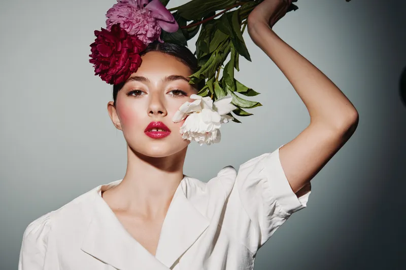 Young woman in white shirt with red and pink flowers in hair poses with arm raised against neutral background. Artistic beauty shot emphasizes natural makeup and floral styling concept.