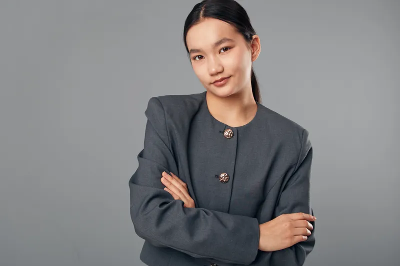 Portrait of confident young woman with crossed arms in gray jacket looking at camera against neutral background. Business style, professional attitude and modern fashion in studio lighting.