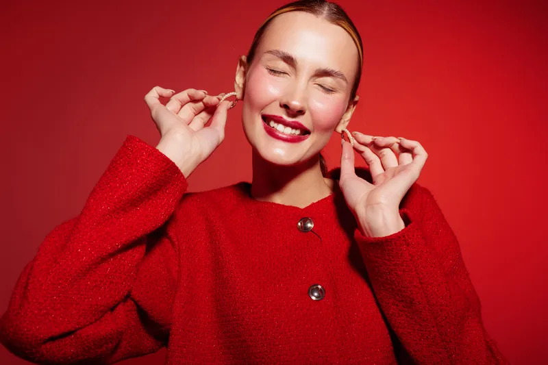 Smiling woman in red coat with closed eyes adjusts earrings on red background. Concept of fashion, beauty, happiness, and style with vibrant color contrast.