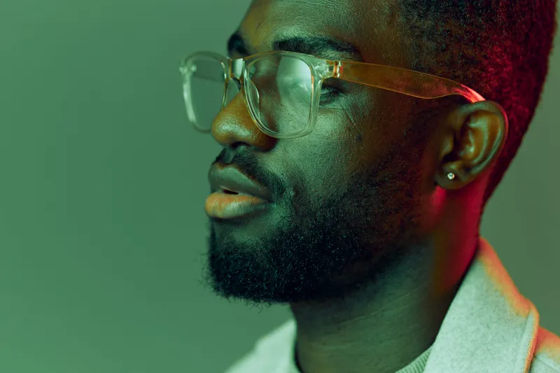 Close up portrait of a young African man with glasses, showcasing confidence against a colorful green background, ideal for concepts of modern style and self expression