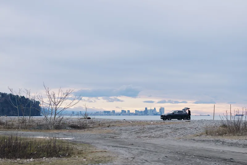 Car on a rocky coast with distant city skyline across calm water at dusk, sparse vegetation and a wide horizon under moody clouds