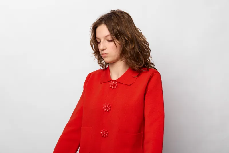 Young woman with curly hair wearing red sweater looks down with serious expression on white background. Concept of sadness, contemplation, and emotional introspection in casual fashion.