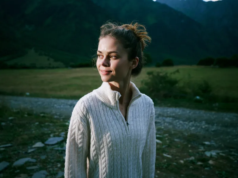 Woman in a cream knit sweater standing outdoors in a mountain valley, serene landscape with soft light, reflective mood, suitable for travel, wellness, or lifestyle themes