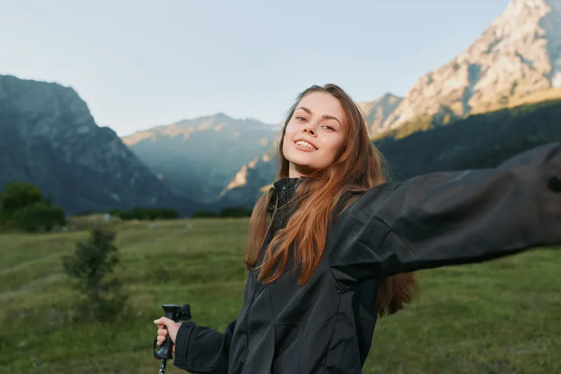 A cheerful outdoor enthusiast enjoys a hike in a scenic mountain valley. She smiles warmly, wearing a weatherproof jacket and gripping a trekking pole, embodying adventure, freedom, and exploration