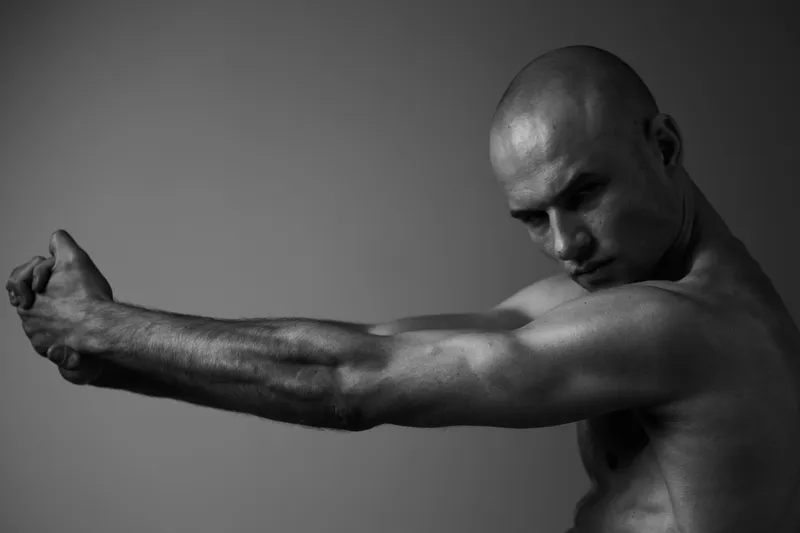 Black and white image of a muscular man stretching his arms, showcasing strength and determination, with a focused expression emphasizing physical fitness.