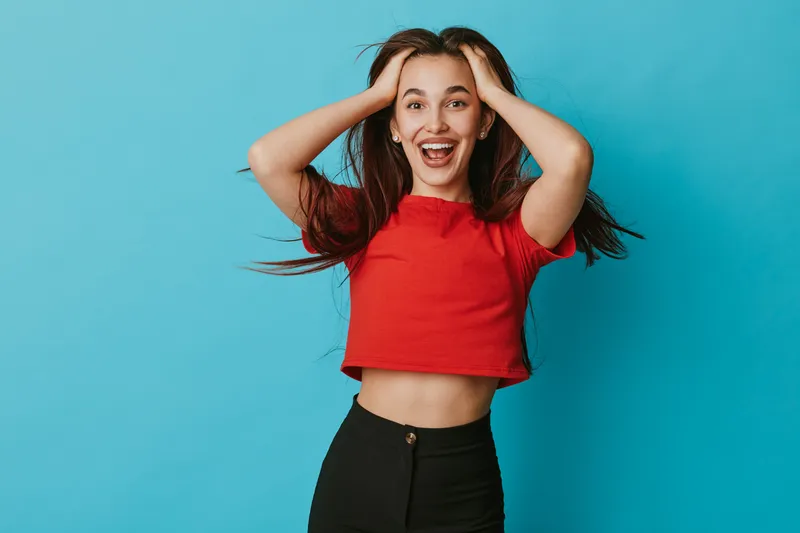 Young woman with long hair and red crop top smiling with hands on head against blue background. Casual style, happiness and excitement in studio portrait with natural expression.
