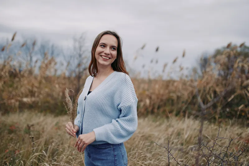 A smiling young woman stands in a windswept meadow holding dried grasses, conveying relaxed genuine presence and natural lifestyle portrait that reflects authenticity and credibility in tone.