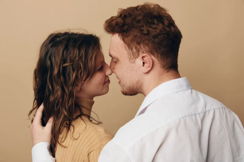 Romantic young couple in casual clothes embraces and touches noses gently, showing affection and intimacy on beige background. Concept of love, connection, and tenderness between partners.