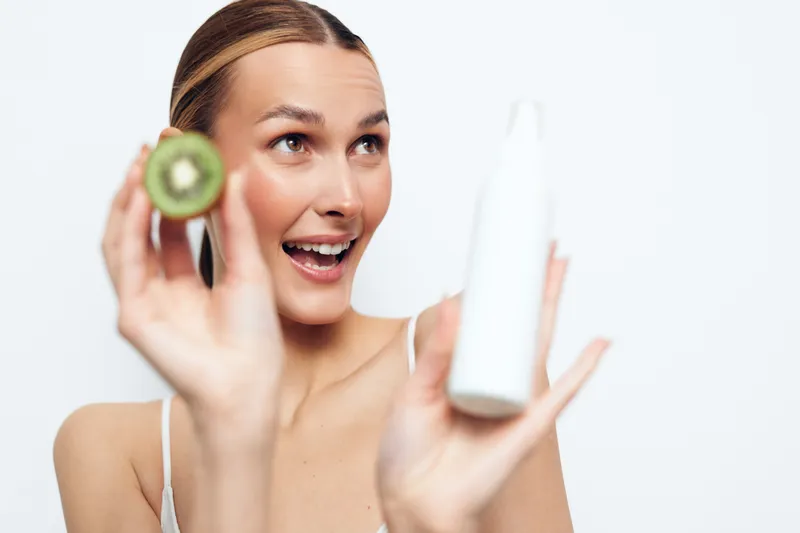 Young brunette woman holds a halved kiwi and a white bottle in a bright studio, smiling during a fresh skincare moment and natural colors, soft lighting, casual pose, copy space nearby.
