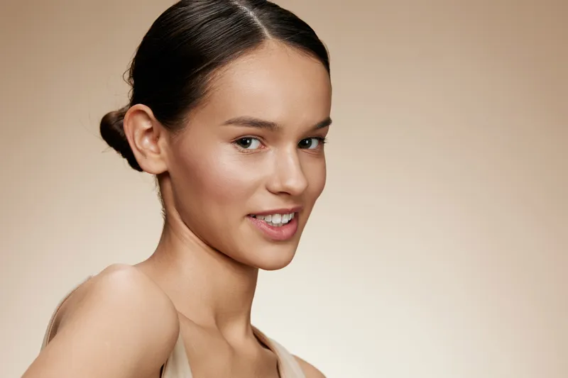 Portrait of a young woman with a natural smile, showcasing healthy skin and minimalist makeup against a soft beige background, perfect for beauty and skincare promotions