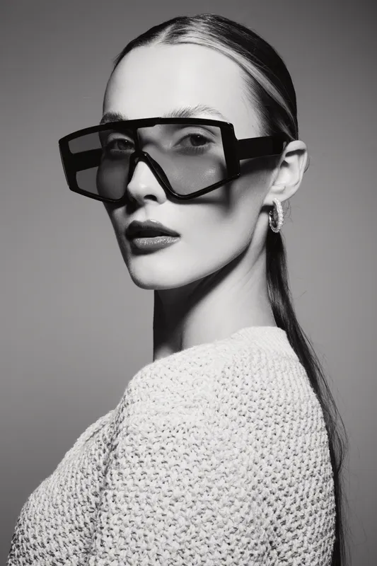 Fashionable young white woman with fair skin wears large geometric sunglasses and textured sweater in monochrome studio portrait. Concept of style and elegance.