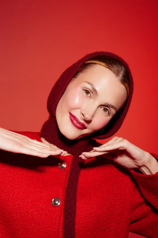Smiling woman in red woolen hood and coat poses with hands near face on red background. Studio portrait with bright colors and warm lighting. Concept of fashion, winter style, and beauty.