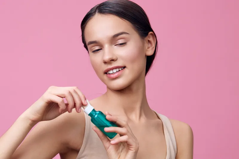 Young woman holding a green serum bottle, showcasing healthy skin and beauty against a soft pink background, exuding confidence and care