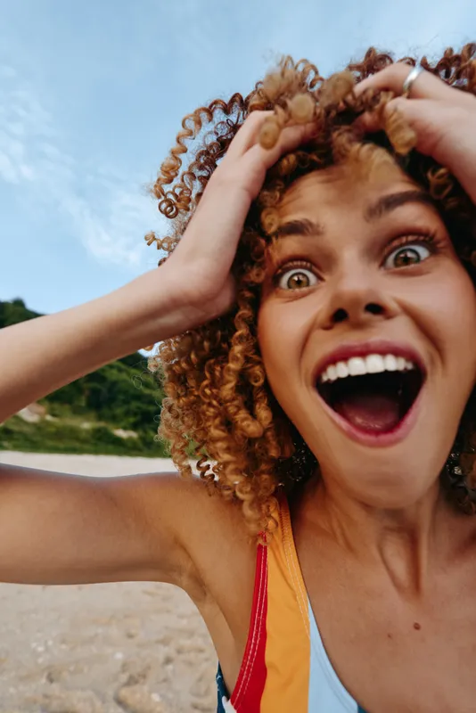 Young woman on a sunny beach laughs with wide eyes and open mouth, curly hair wild and hands framing her face, capturing a spontaneous joyful moment outdoors