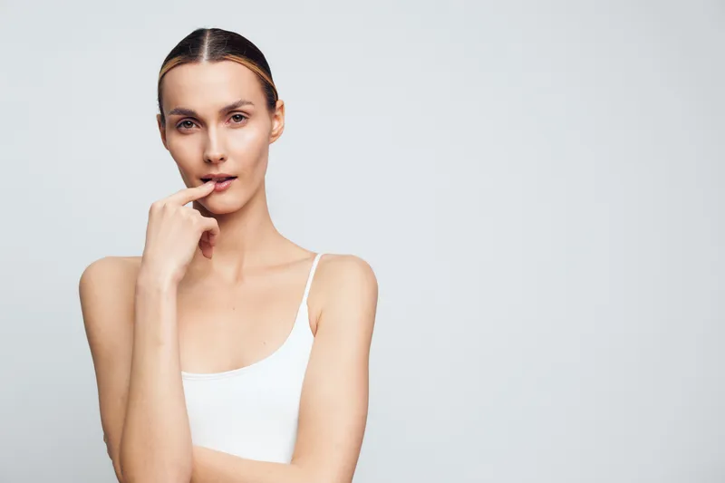 Young woman with clear skin wearing a white tank top posing with finger near lips on a light gray background. Concept of beauty, skincare, minimalism, and natural look.