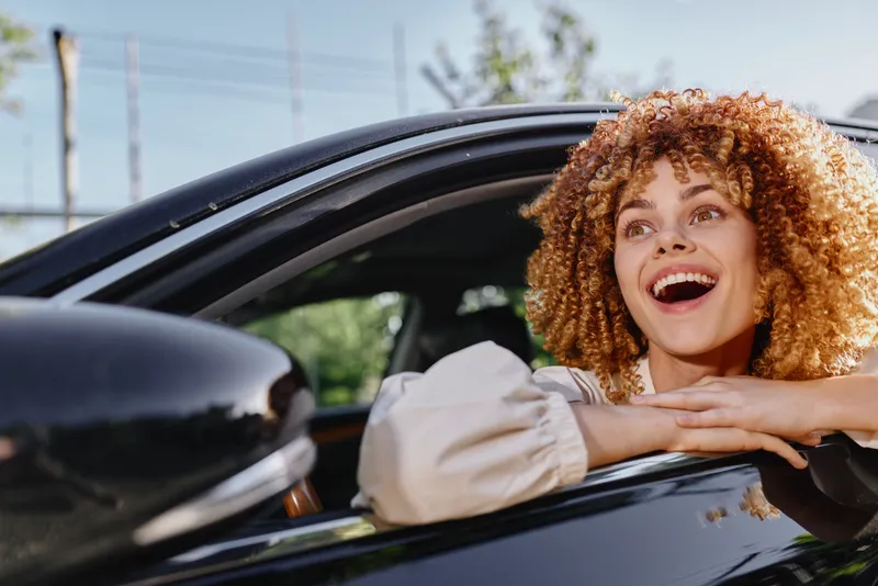 curly woman looking excitedly out of car window, sunny day, casual beige blouse, outdoor, joyful expression, natural light