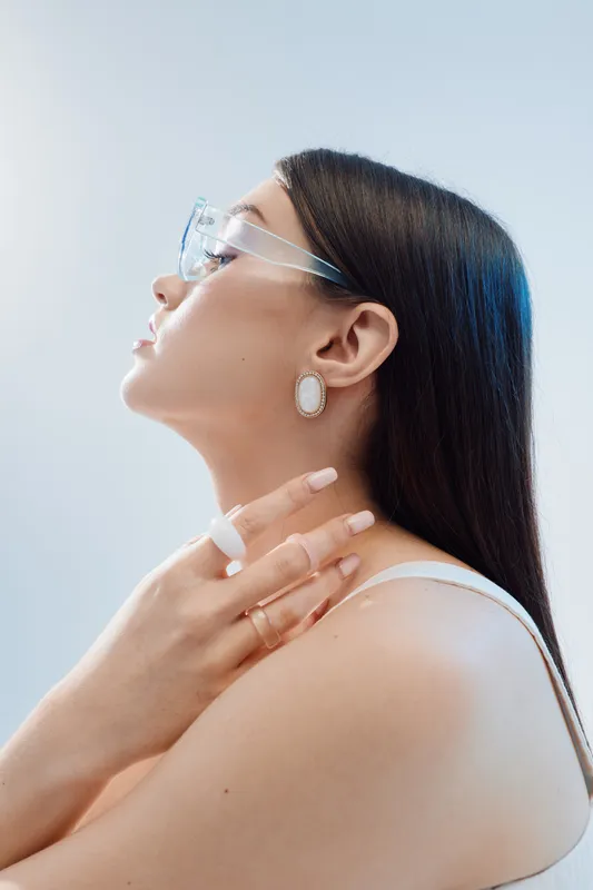 Elegant woman with long dark hair wearing transparent glasses and white earrings poses in profile. Beauty fashion concept with minimalistic style and soft lighting.