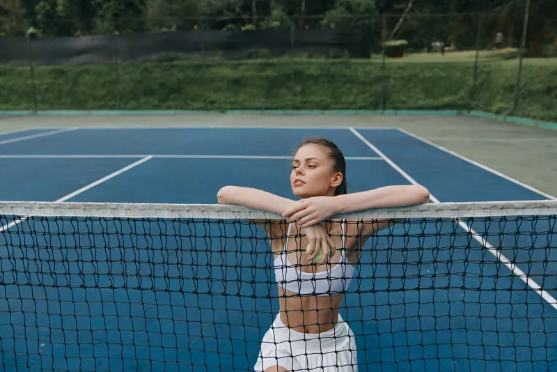 Athletic young woman resting on tennis net during a sunny day on the court, showcasing sports fashion and a focused expression Key elements include tennis, fitness, and summer