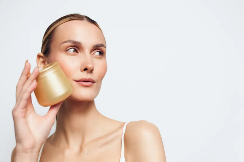 Young woman with clear skin holds golden cosmetic jar near face looks aside with calm expression isolated on white background beauty skincare concept.