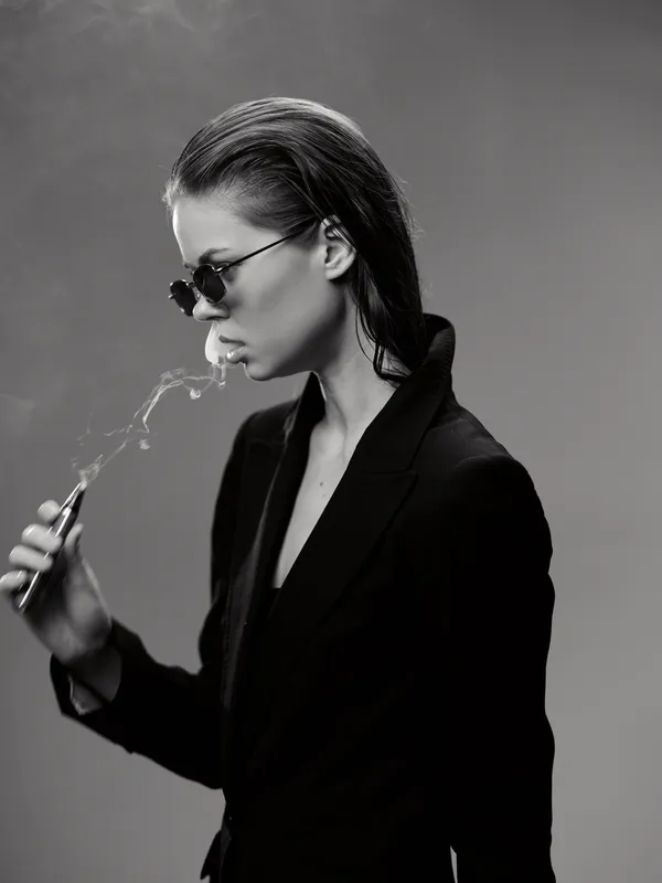 Black and white portrait of a stylish woman smoking with sunglasses and sleek hair in elegant dark clothing
