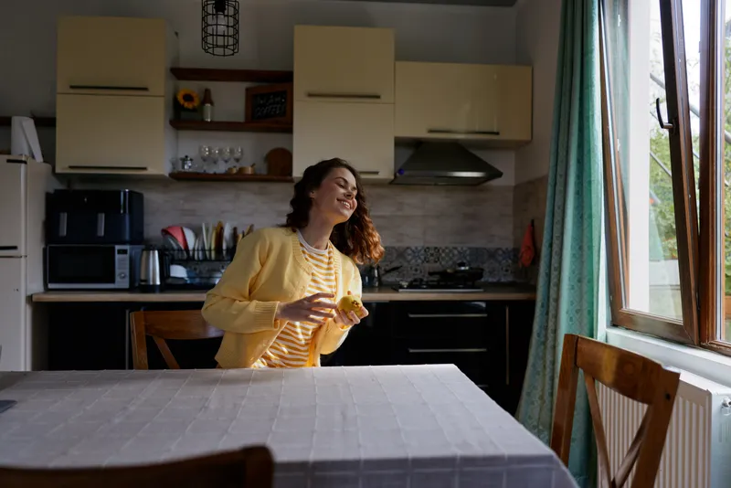 A cheerful woman in a yellow cardigan stands at a kitchen table, smiling, as sunlight pours through a window. A cozy home scene highlighting warm everyday moments.