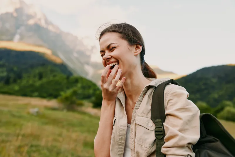 Happy woman with backpack laughing outdoors in nature with mountains in the background. Concept of lifestyle, travel, adventure, and freedom in natural light.
