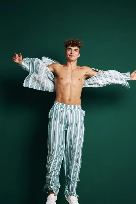 Young man with striped pajama pants and matching open shirt smiling and spreading arms against dark background for casual cozy style and vibrant mood in relaxed standing pose.