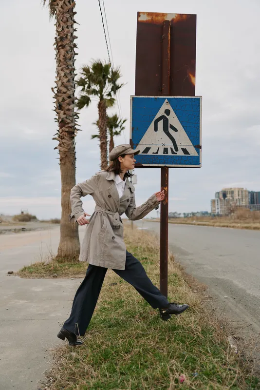 Woman in trench coat and hat interacts with pedestrian crossing sign outdoors near palm trees. Urban environment with road and buildings in background. Concept of street style and city life.