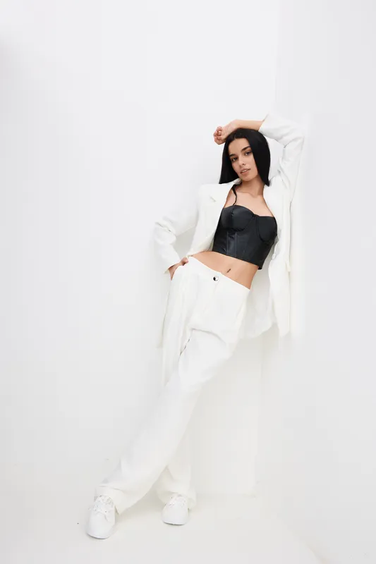 Stylish woman in white pants and blazer posing confidently against a clean white background with a black top, showcasing modern fashion and elegance.