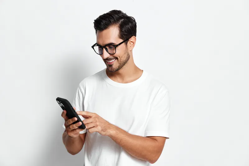 Smiling man with glasses wearing a white t-shirt, looking at a mobile phone while standing against a plain solid background. Casual technology lifestyle concept.