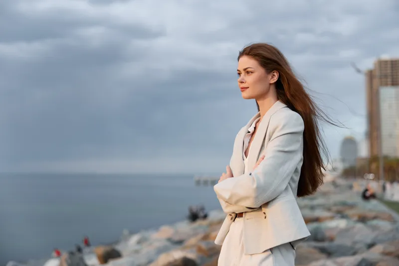 Confident woman in business attire enjoying a serene coastal view, showcasing elegance and professional demeanor amidst a muted color palette.