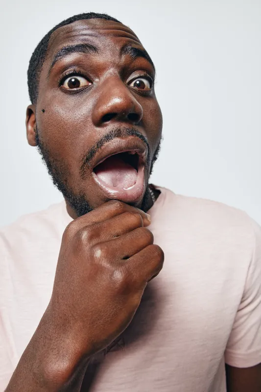 African American man with dark skin and short hair showing surprised expression with open mouth and hand on chin against white background. Casual clothing and emotional reaction.