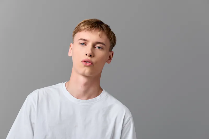 Man in a white shirt poses casually with a subtle pout against a gray studio backdrop, conveying a relaxed, modern lifestyle with soft lighting and minimal styling