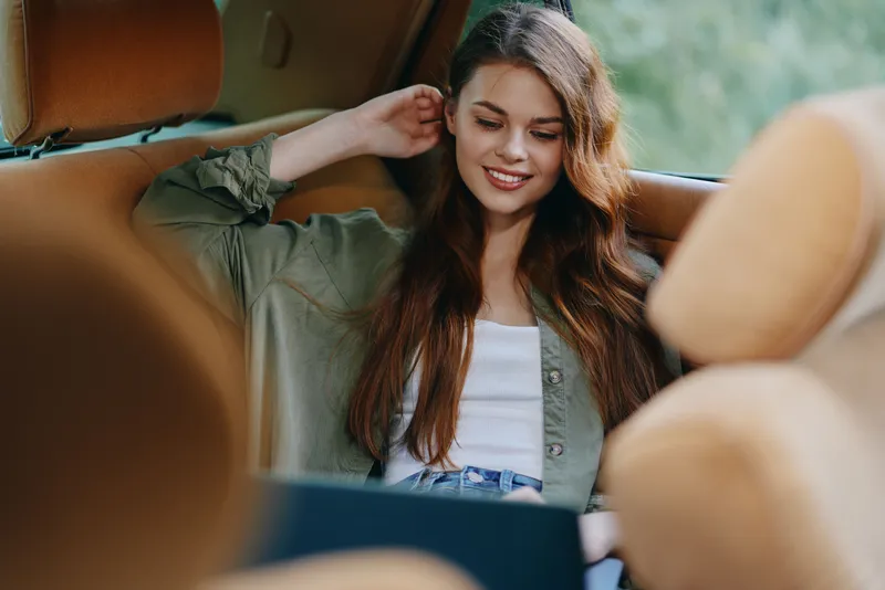 Young woman enjoying her time in a car with a laptop, displaying a joyful expression, dressed casually in a green shirt, perfect for lifestyle and travel concepts.