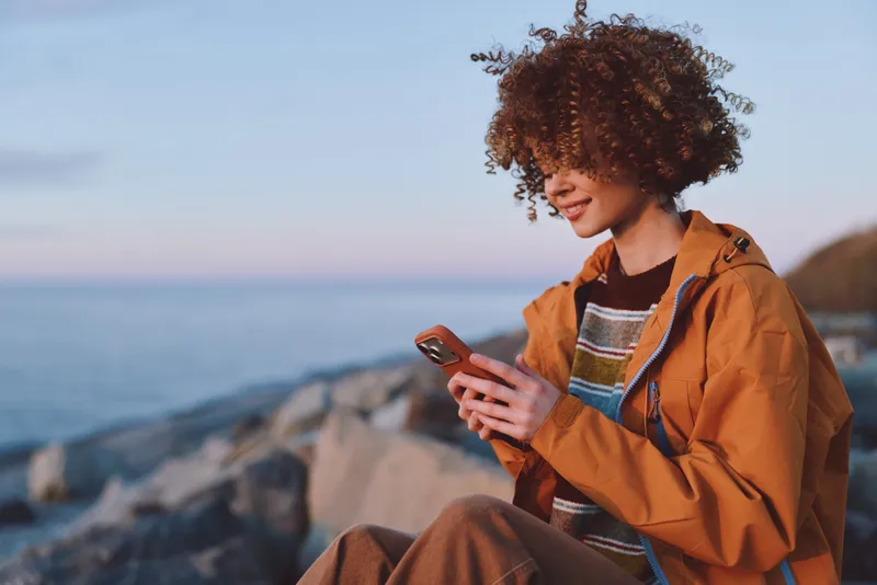 Woman lifestyle portrait outdoors by sea, sitting on rocky coast, curly hair, orange jacket, looking at smartphone, relaxed candid mood, travel concept, sunrise daylight