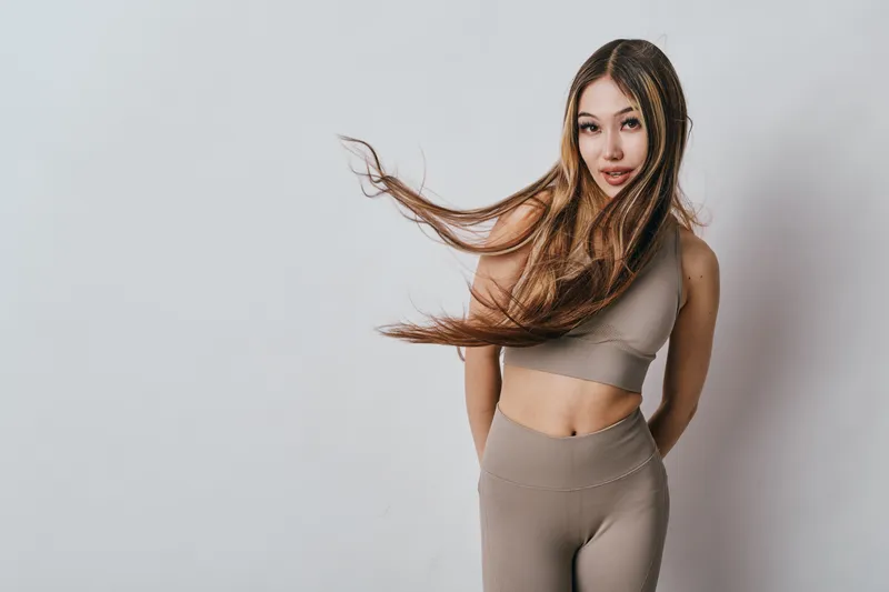 Young woman with long hair wearing beige sportswear posing against white background. Fitness model standing with flowing hair and casual expression in studio. Healthy lifestyle concept.