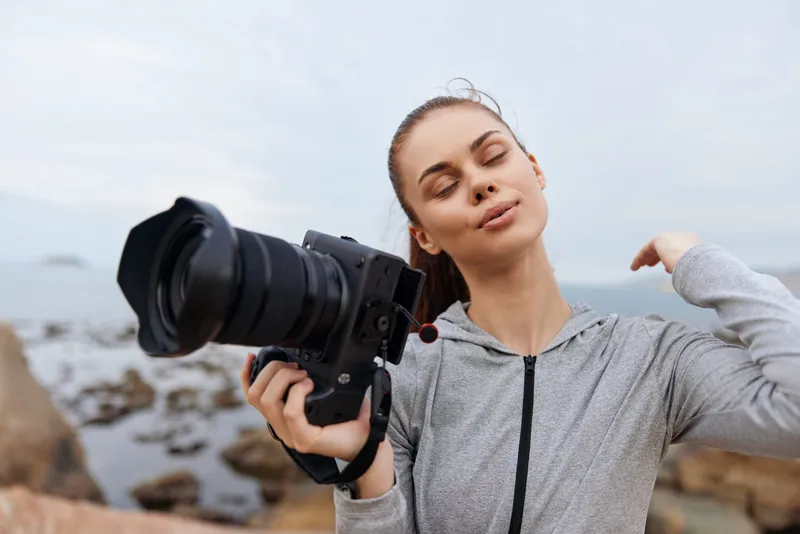 Young woman confidently holding a camera, enjoying the outdoor scenery by the sea, showcasing a casual and trendy look against a natural background
